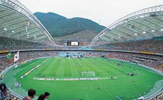 A crowded soccer stadium with arched roofs overlooks a match, with a green mountain in the background.