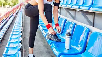 Woman in workout clothes ties her shoelace, foot on a bright blue stadium seat beside a water bottle.