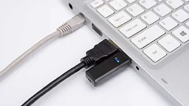 Close-up of a silver laptop with a grey Ethernet cable and a black HDMI adapter, featuring a blue indicator light, plugged into its side ports.