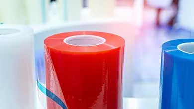 Three large, upright rolls of shiny plastic film in white, red, and blue, against a blurred, brightly lit background.