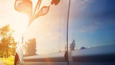Close-up of a car's side with sunlight flaring over the side mirror and blue sky reflected in the window.