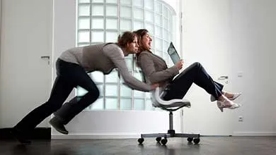 Two women in an office laugh as one pushes the other, who is holding a laptop, in a rolling chair.