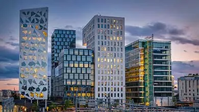 City skyline at dusk with a prominent white building featuring geometric cutouts, surrounded by illuminated glass and concrete high-rises beneath a purple sky.