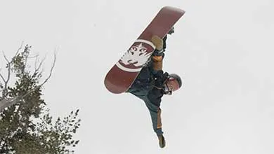 A snowboarder hangs upside down in mid-air, gripping the edge of a maroon snowboard against a cloudy sky.