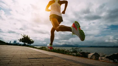 Low-angle rear view of a person running on a paved path beside water, mid-stride with one leg extended back. Sunlight breaks through clouds, and a city skyline appears in the distance.