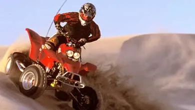 An all-terrain vehicle riding over sand dunes, kicking up sand under a clear blue sky.