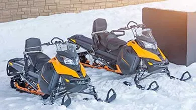 Two black and yellow snowmobiles parked side by side on snow next to a brick wall.