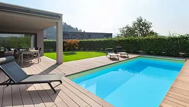 Modern residential backyard with a rectangular swimming pool, wood deck, lounge chairs, and landscaped greenery.