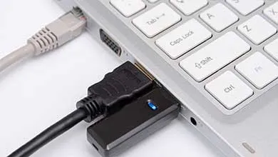 Close-up of a silver laptop showing part of its white keyboard with visible Tab, Caps Lock, Shift, and Ctrl keys. Three cables are connected: a gray Ethernet cable, a black HDMI cable, and a black USB device with a blue indicator light.