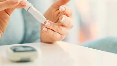 Hands holding an auto‑injector pen while adjusting or preparing the device, with a glucose meter resting on a tabletop in the foreground.