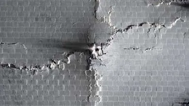 Close-up of damaged gray drywall with intersecting cracks and peeling surface over a textured grid pattern.