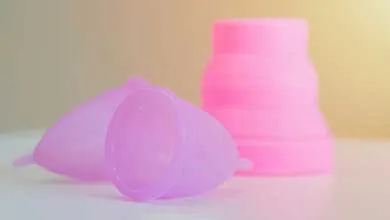 Two translucent lavender menstrual cups lie on a white surface in the foreground, with a pink collapsible silicone cup softly blurred against a cream background.